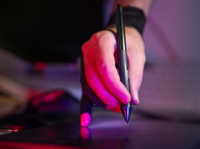 A person holding a pen and working on a laptop, creating content for an artist’s guide to selling.