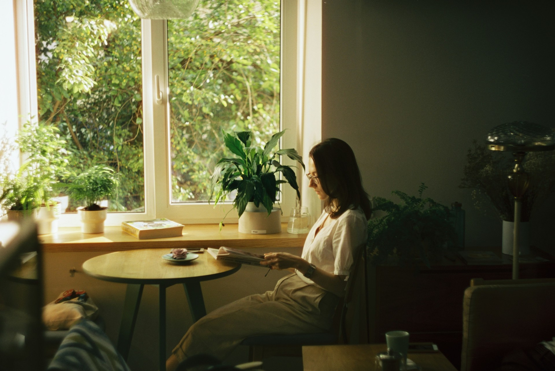 A woman reading at a table surrounded by plants, deep in thought as her inner critic lingers in the background.