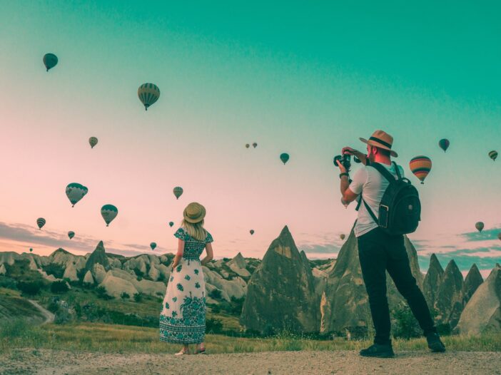 A man photographing hot air balloons in the sky while a woman stands in front of him, embodying the spirit of travel journalism.